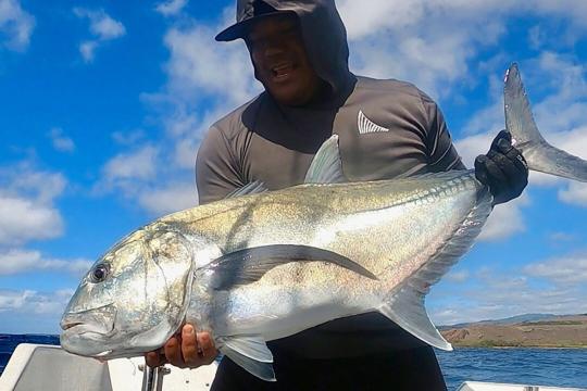 A smiling fisherman holds a nearly 35-inch-long silvery fish in his hands while on a boat in the ocean.