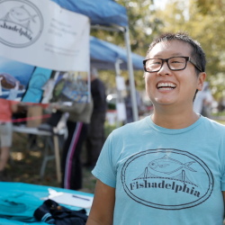 A Chinese-American woman with short hair and glasses wears a blue Fishadelphia t-shirt at a fish stand.