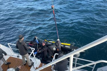 The view looking down to a lower deck of a ship and the ocean from an upper deck. An inflatable boat is alongside the ship, and a yellow glider in the water on the other side of the inflatable boat. Several people are in the boat and on the ship, attaching a rope to the glider so it can be lifted out of the water. 