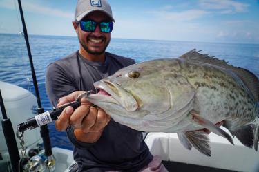 A white man in sunglasses and a baseball cap smiles and holds a fish up to the camera.