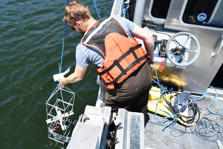 A man wearing an orange lifejacket lowers scientific equipment over the side of a boat.